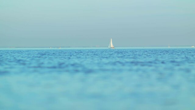 Beautiful little white sailboat yacht with white sail anchored in the deep blue sea away from the coast at evening sunset