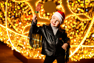 a beautiful, cheerful little girl in a Santa Claus hat,standing near the lights,with a lollipop in her hand on a festive evening, on the street