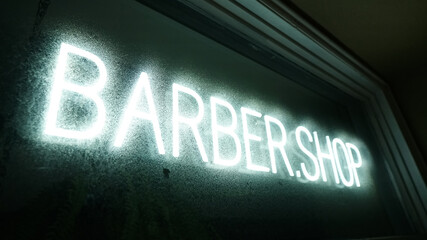 Illuminated neon white sign that spells BARBERSHOP displayed behind a condensed window glass plane. 