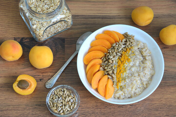 cooked oatmeal with apricot slices, sunflower seeds, curcuma in white plate with teaspoon