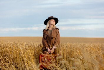 Blonde woman in cloak and hat with suitcase in wheat field