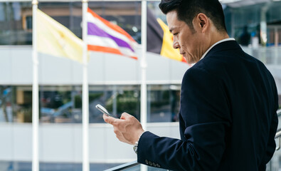 Serious Asian Businessman in Blue Suit Using Mobile Phone while Standing at Modern Office Building with Thailand and Belgium flag in the Background