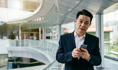 Naklejka na ściany i meble Portrait of Handsome Asian Businessman Using Mobile Phone and Standing at Modern Balcony Outdoors 