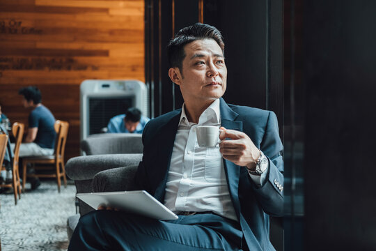 Handsome Asian Businessman Using Digital Tablet in a Cafe.
Serious business man in blue suit working on a digital tablet while sitting in a restaurant and drinking cup of coffee. - Powered by Adobe