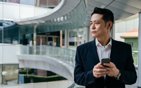 Portrait of Handsome Asian Businessman Using Mobile Phone Outside Office. 

Serious business man in blue suit standing at modern balcony and typing text message on his smartphone while looking away.