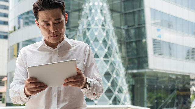 Confident Asian Businessman Using Digital Tablet Outdoors. 

Serious business man watching something on digital tablet while standing at city street with office building in the background.