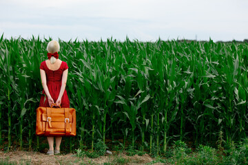 Blonde lady in beautiful red dress with suitcase in cornfield