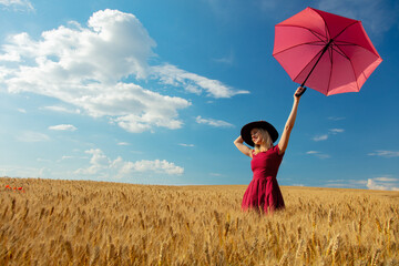 Blonde girl in hat and red dress with umbrella in wheat field