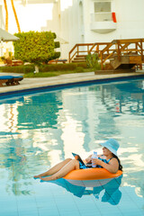 A woman in a sun hat in the pool in a swimming circle with a laptop works remotely on the background of sun loungers.