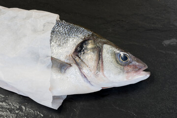 Fresh Sea bass wrapped in white paper, fresh from the market on black stone background