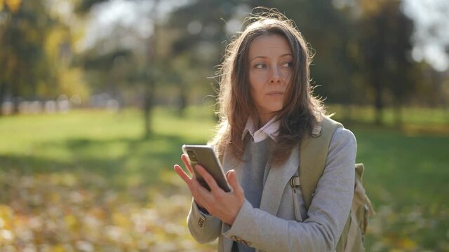 A pretty adult female tourist with a phone and a backpack is standing in a city park and trying to find a map on the Internet to search for routes