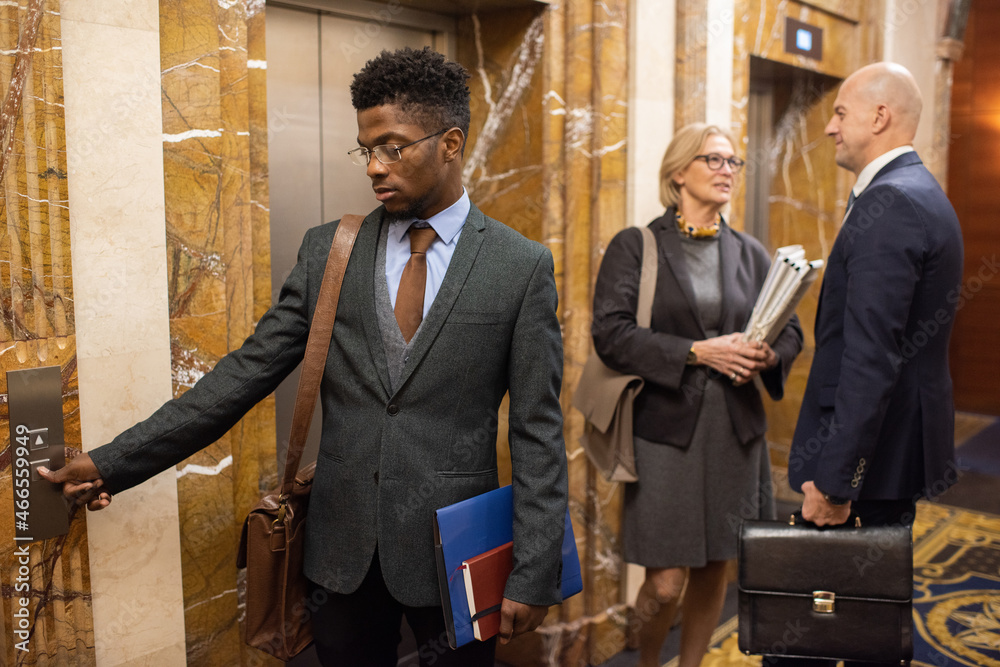 Poster young elegant businessman pushing elevator call button while standing against two co-workers inside 
