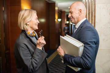 Two elegant mature colleagues discussing news or working points while standing in corridor of...