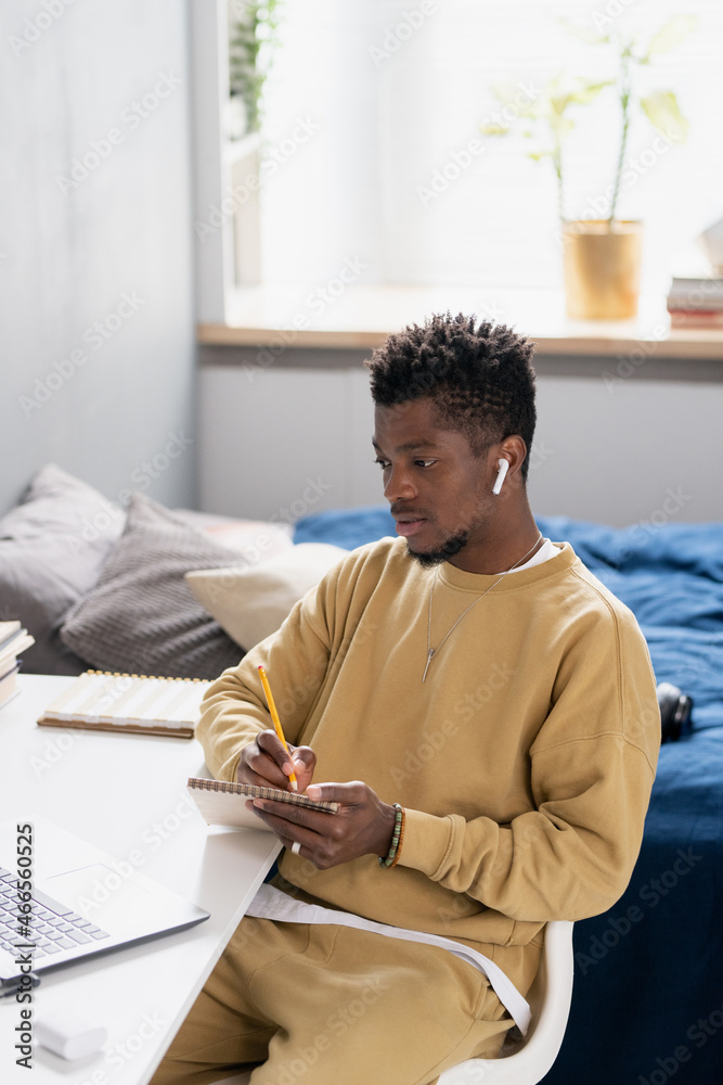 Poster serious young african student making notes in notepad while sitting by desks in front of computer