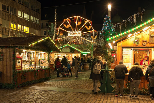 Freiburg im Breisgau, Germany. Small Christmas market at Kartoffelmarkt (Potato Market) square in night. Unknown people consume street food and sweets close to market stalls.