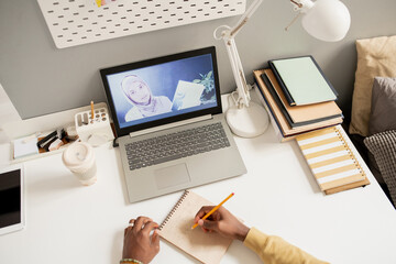 Young confident teacher in hijab on laptop screen and hands of African student making notes during online lesson