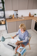 Mature female student communicating with teacher during online course in front of laptop while sitting by kitchen table