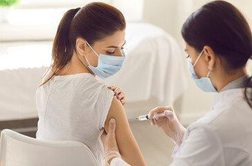Young woman wearing mouth covering getting Covid 19 shot in arm at new modern vaccination center. Nurse in white gloves giving preventive vaccine injection to patient during seasonal flu outbreak - obrazy, fototapety, plakaty