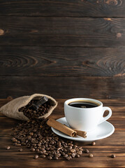 A white ceramic cup with black aromatic coffee on a dark wooden table. Cinnamon tubes on a saucer. Canvas bag with roasted coffee beans in the background