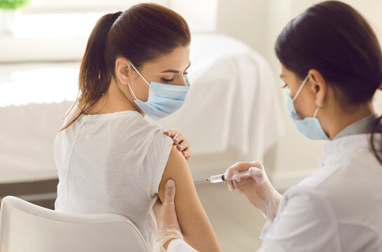 Young Woman Wearing Mouth Covering Getting Covid 19 Shot In Arm At New Modern Vaccination Center. Nurse In White Gloves Giving Preventive Vaccine Injection To Patient During Seasonal Flu Outbreak