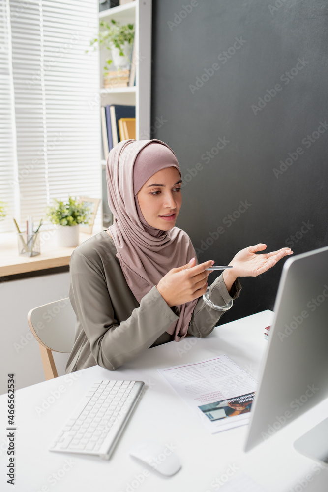 Poster young confident teacher in hijab speaking to online audience in front of computer monitor while sitt