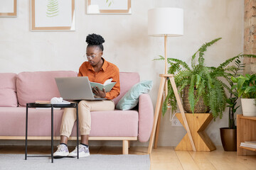 AFrican girl in casualwear sitting on couch against wall in living-room and making notes during online lesson