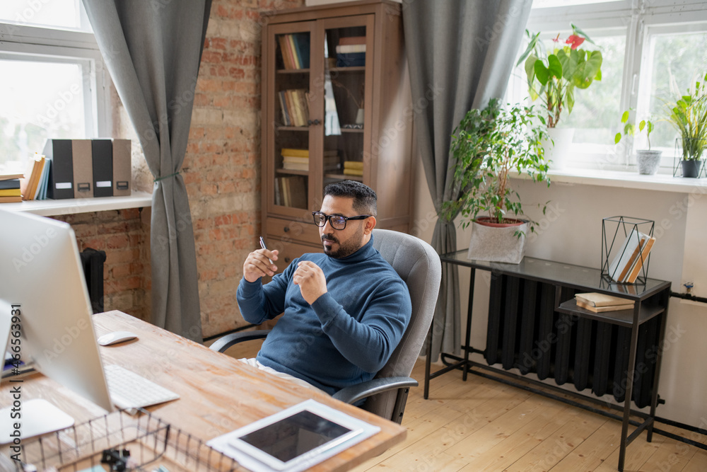 Poster serious teacher in casualwear listening to online student while sitting in armchair in front of comp