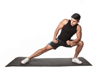 Portrait of a young adult athletic man in sports clothing doing leg stretching exercises on yoga mat before workout, healthy lifestyle. Indoor studio shot isolated on white background.