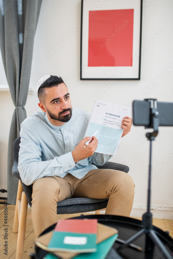 Poster young confident man pointing at paper and looking at smartphone camera while explaining information 