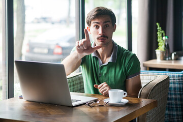 Portrait of man freelancer wearing green T-shirt, sitting and looking at camera with loser gesture, sitting in front of laptop, feels sad. Indoor shot near big window, cafe background.
