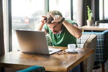 Portrait of angry businessman wearing green T-shirt at work, looking at laptop monitor, showing...