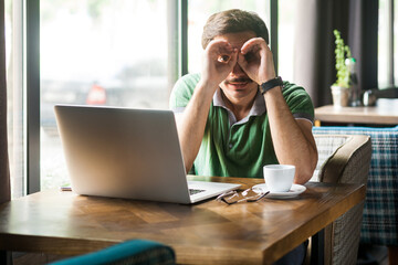 Portrait of businessman wearing green T-shirt, working online on laptop, looking through fingers...