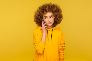 Fototapeta na wymiar Serious woman with Afro hairstyle using mobile phone, making call and talking with friend, connection, wearing casual style hoodie. Indoor studio shot isolated on yellow background.
