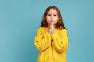 Portrait of little girl praying to God apologizing for bad behavior, looking with imploring eyes, wearing yellow casual style sweater. Indoor studio shot isolated on blue background. - obrazy, fototapety, plakaty