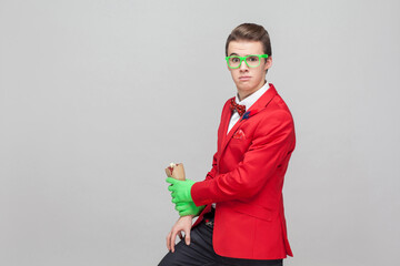 Portrait of stylish bizarre gentleman with eyeglasses in red tuxedo and green gloves holding zombie...