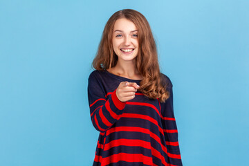 Hey you! Happy woman wearing striped casual style sweater pointing to camera and looking with charming toothy smile, indicating finger, making choice. Indoor studio shot isolated on blue background.