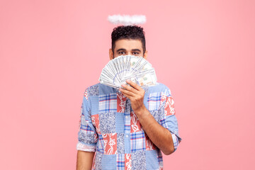 Angelic rich bearded man wearing blue casual style shirt with nimbus on head peeking out of dollar banknotes, rejoicing big profit earn. Indoor studio shot isolated on pink background.