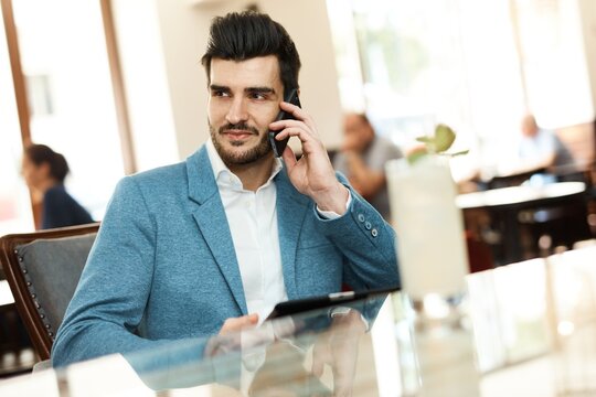 Young businessman sitting at cafeteria, using mobilephone and tablet, looking away.