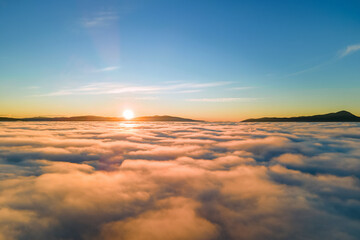Aerial view of vibrant sunset over white dense clouds with distant dark mountains on horizon.