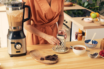 Hand of young woman grating dark chocolate for tasty homemade cocoa dessert or smoothie in electric blender
