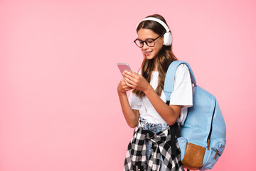 Cropped shot of a caucasian young girl teenager schoolgirl pupil using smart phone while listening to the music in headphones going back to school isolated on pink background.