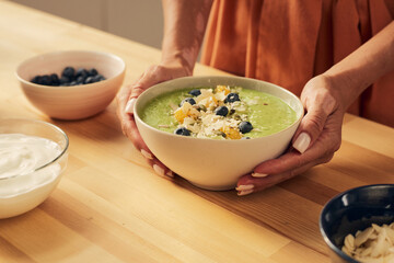 Hands of woman holding bowl with fresh homemade smoothie decorated with blueberries, muesli, oranges and almond flakes