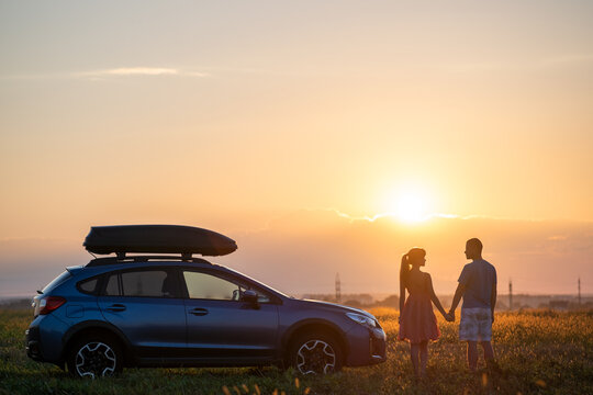 Happy couple standing beside their SUV car during honeymoon road trip at warm summer evening. Young man and woman enjoying time together travelling by vehicle.