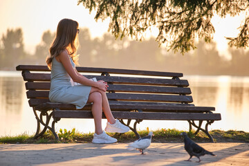 Lonely woman sitting on lake side bench enjoying warm summer evening. Solitude and relaxation concept