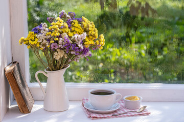 Fototapeta na wymiar Old book, colorful bouquet of flowers and white cup of tea on background of window with raindrops at home at summer day