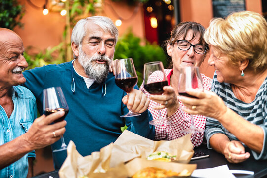 Happy seniors having fun toasting red wine at dinner garden party - Retired couples drinking at restaurant together - Dinning friendship concept on warm vivid filter - Focus on bearded hipster man