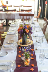 Table decorations at a wedding follow a purple and orange colored theme. The center pieces include flower petals, a wine bottle and purple sash going across the table.