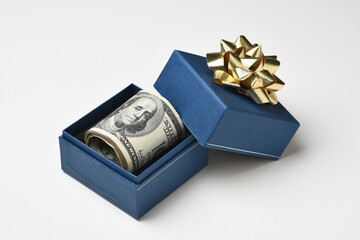 A blue gift box and gold bow with a roll of one hundred dollar bills.