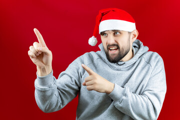 against a Christmas red background, a man in a red Santa Claus hat is standing and pointing with his forefingers to the side.