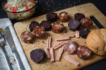 small pieces of sausage on the wooden board with spices. Cabbage salad in a plate with a silver fork and spoon, black background